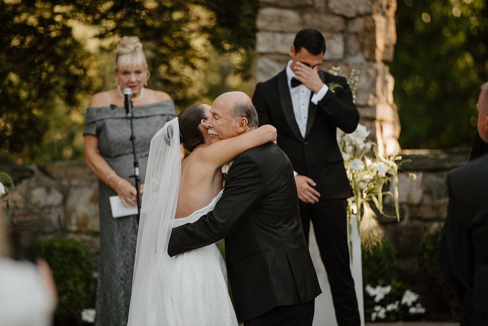 Bride hugging her father at the altar while the emotional groom wipes away a tear in the background.